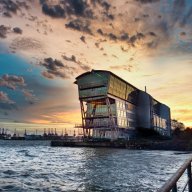 Das Alte Fährterminal bei Sonnenuntergang, aus Richtung der Hamburger Innenstadt fotografiert. Im Vordergrund ist das Wasser der Elbe zu sehen, links im Hintergrund die Kräne des Hafens.