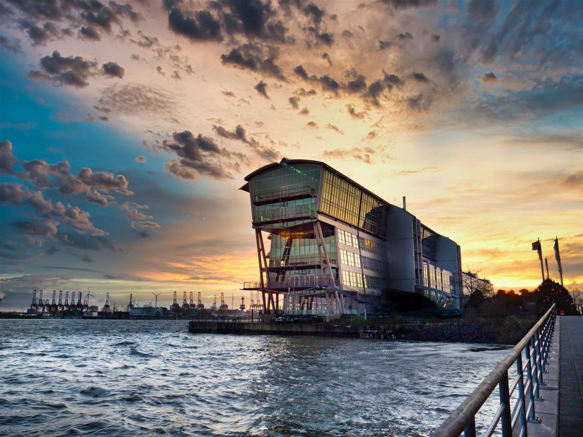 Das Alte Fährterminal bei Sonnenuntergang, aus Richtung der Hamburger Innenstadt fotografiert. Im Vordergrund ist das Wasser der Elbe zu sehen, links im Hintergrund die Kräne des Hafens.