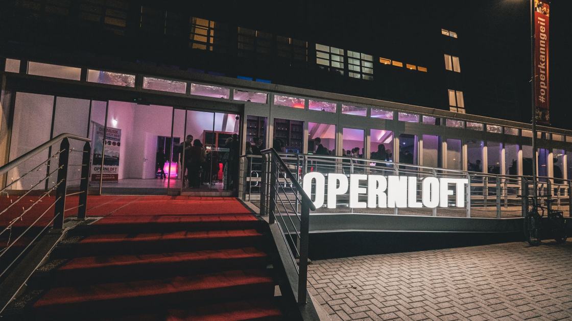 Vorderansicht des Opernlofts bei Dunkelheit. Ein roter Teppich liegt auf der Treppe und der Schriftzug des Opernlofts, welcher vor der Fassade steht, leuchtet. 
