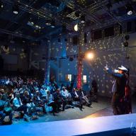 Ein Sänger und eine Sängerin stehen auf der Bühne und tragen Kapitänsmützen. Die Perspektive ist auf das Publikum gerichtet, Eine Diskokugel wirft Licht durch den Raum. 