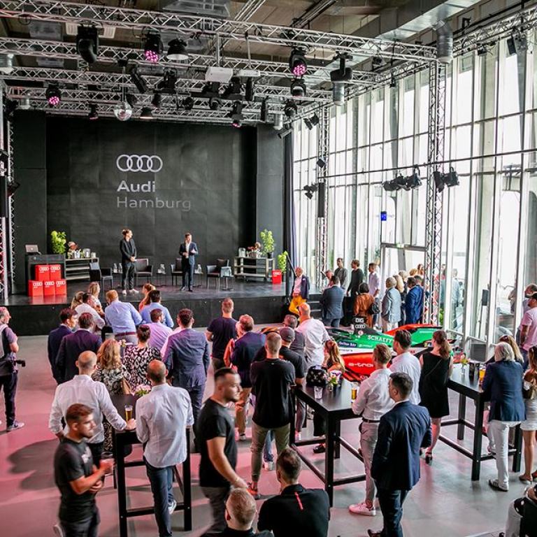 Eine Veranstaltung von Audi, zwei Redner stehen auf der Bühne, im Publikum sind etwa 30 Personen zu sehen. Teile eines Audi Formula E Wagens sind zu sehen.