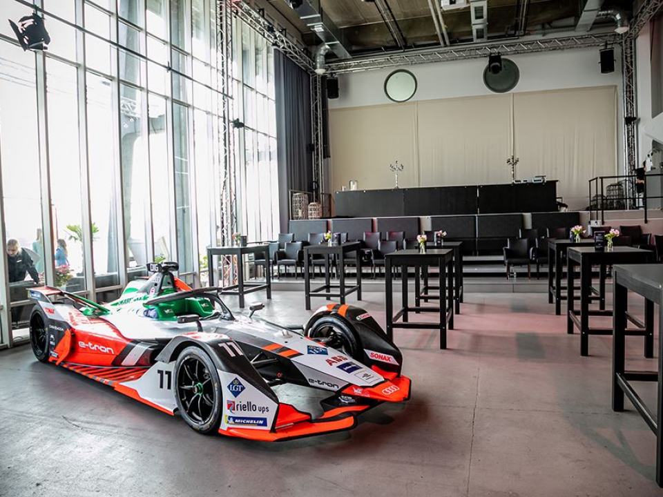 Der Saal bei einer Vernstaltung von Audi-Sport. im Vordegrund steht ein Audi Formula E Rennwagen. Die Bestuhlung des Saals besteht aus Hochtischen und wenigen Reihen mit Bequemen Lederstühlen.