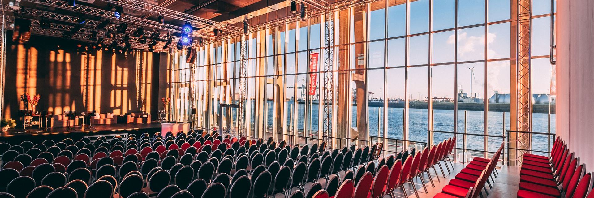 Der Saal des Opernlofts in normaler Bestuhlung mit frontaler Bühne. Auf der rechten Seite des Saals kann man durch die Fensterfront den Hafen sehen.