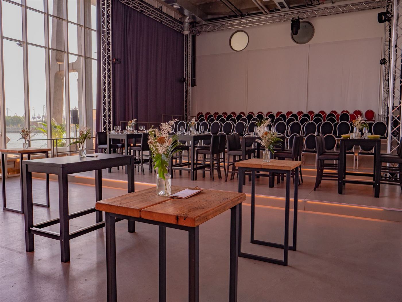 Alternative Bestuhlung im Saal des Opernofts. Im Hintergrund ist eine kleine Tribühne mit 7 Reihen, im Vordergrund stehen einige Hochtische mit sommerlichen Blumensträußen in Glasvasen.