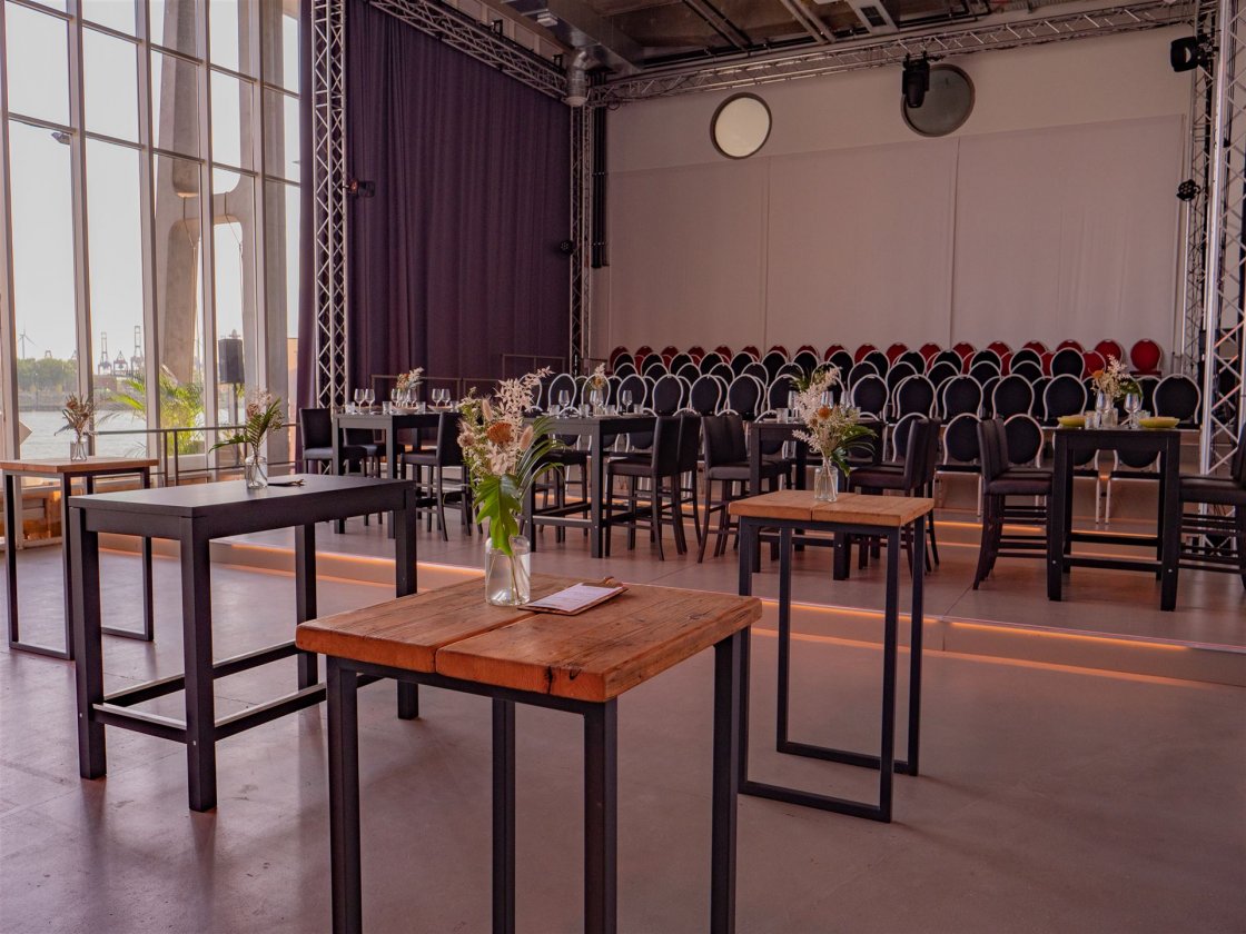 Alternative Bestuhlung im Saal des Opernofts. Im Hintergrund ist eine kleine Tribühne mit 7 Reihen, im Vordergrund stehen einige Hochtische mit sommerlichen Blumensträußen in Glasvasen.