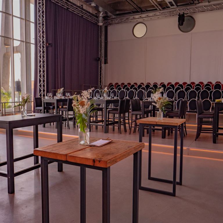 Alternative Bestuhlung im Saal des Opernofts. Im Hintergrund ist eine kleine Tribühne mit 7 Reihen, im Vordergrund stehen einige Hochtische mit sommerlichen Blumensträußen in Glasvasen.