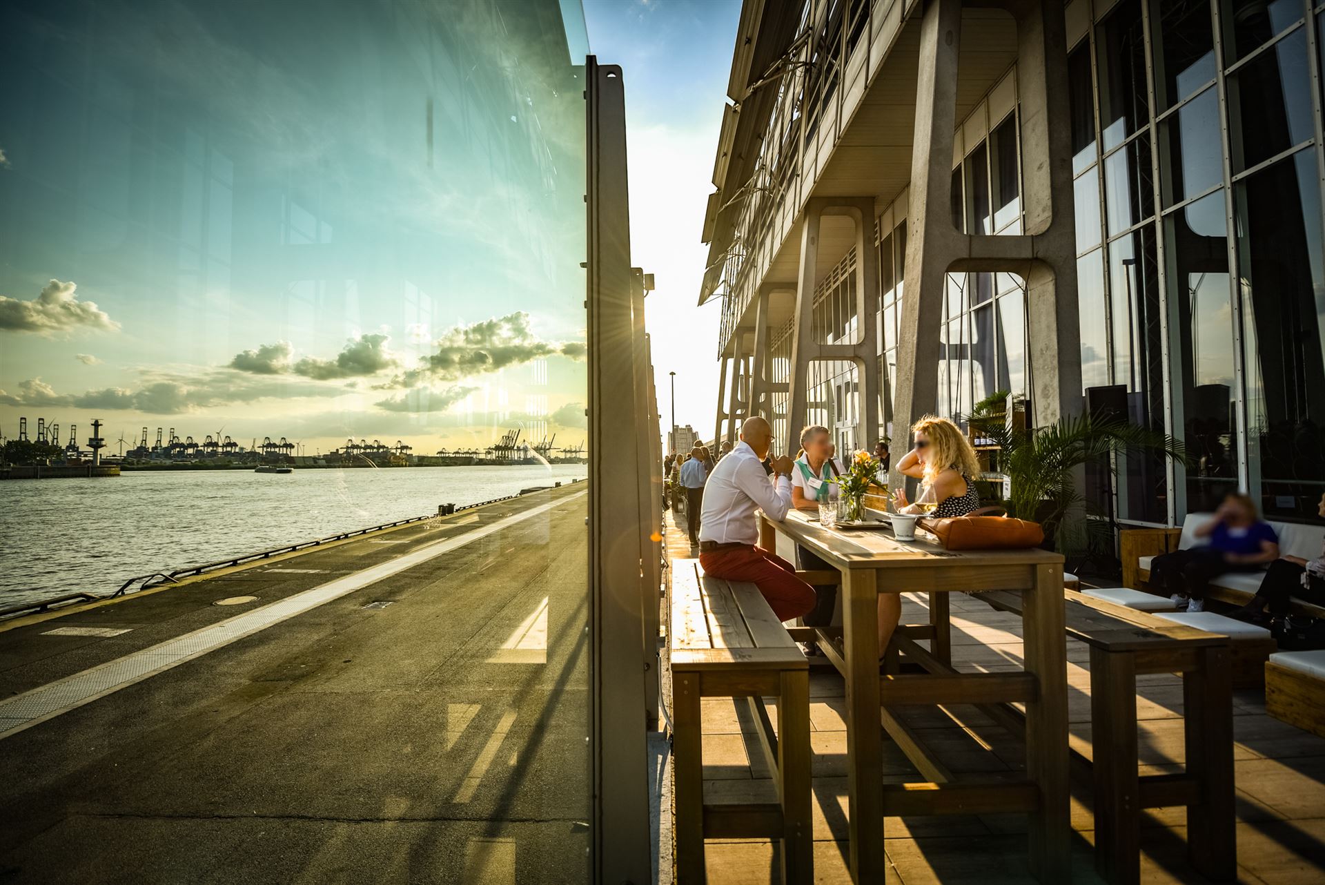 Teilnehmer einer Veranstaltung auf der Elbterasse im Sommer. Im Vordergrund sitzen drei Personen an einem Hochtisch.