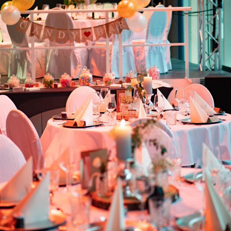 Der Saal ist für eine Hochzeit dekoriert. Zu sehen sind einige eingedeckte Rundtische.