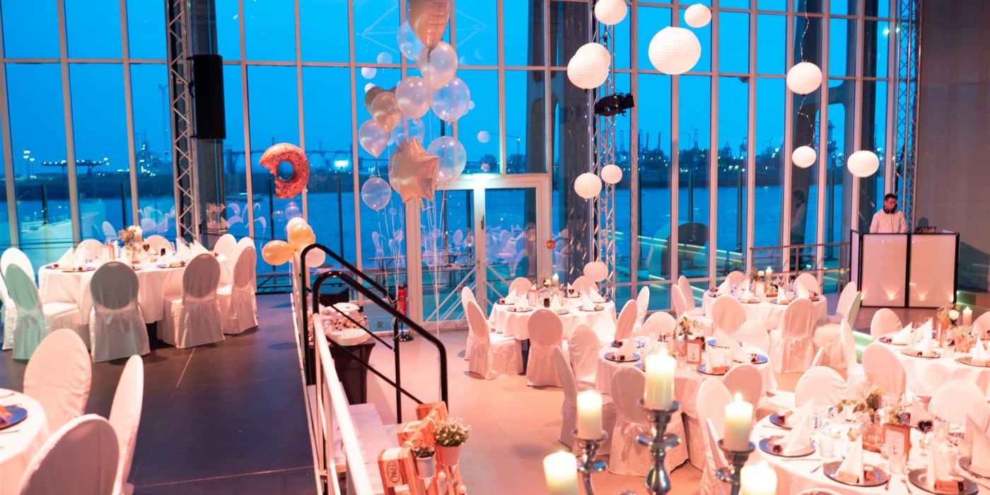 Der Saal ist für eine Hochzeit dekoriert. Im Vordergrund sieht man einen Kronläuchter und im Hintergrund Ballons und weiße Lampignons, die an der Decke hängen. im Saal stehen viele Rundtische.